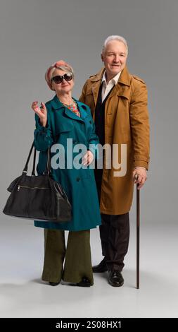 Couple en trench-coat, femme regardant loin avec un sourire réfléchi, homme debout avec canne et regard chaud et élégance sur fond gris. Banque D'Images