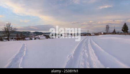 Petit village alpin et hiver montagne enneigée au premier lever du soleil autour, Voronenko, Carpates, Ukraine. Tracer sur un chemin fraîchement trodden à travers g Banque D'Images