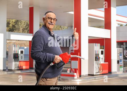 Homme mature souriant tenant un pistolet de ravitaillement et faisant des gestes avec les pouces vers le haut à une station-service Banque D'Images