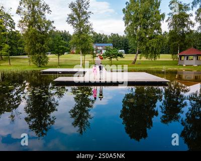 Deux enfants se tiennent debout sur un quai en bois au-dessus de l'eau calme, reflétant les arbres et les nuages. Une maison de toit rouge et d'autres bâtiments sont visibles parmi la verdure luxuriante Banque D'Images