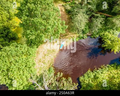 Une perspective aérienne montre une planche de paddleboard sur une rivière sinueuse entourée d'une forêt dense et verdoyante, mettant en évidence le contraste entre feuillage et eau. Banque D'Images