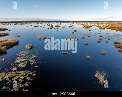 Une vaste zone humide présente une grande étendue d'eau avec des îlots herbeux. Un bateau isolé crée des ondulations, tandis que le ciel dégagé et les nuages dispersés se reflètent sur le Banque D'Images