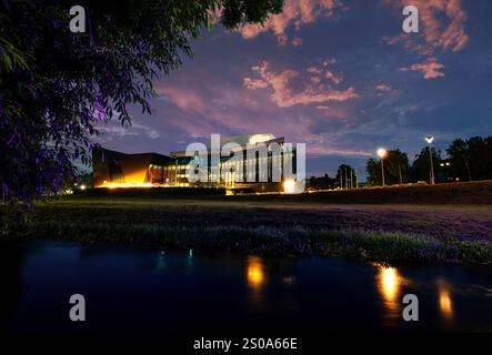 Un bâtiment moderne à Rezekne, en Lettonie, avec de grandes fenêtres en verre, est illuminé contre un ciel nocturne vibrant, reflétant sur un plan d'eau calme. Banque D'Images