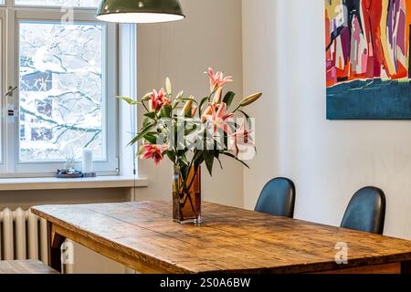 Une salle à manger confortable dispose d'une table en bois avec des lys roses, éclairée par une lampe suspendue. Les branches couvertes de neige sont visibles à travers la fenêtre. Banque D'Images
