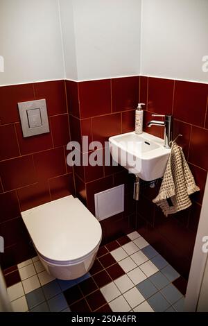 Une salle de bains compacte est dotée de murs carrelés d'un rouge profond, de toilettes murales et d'un petit lavabo d'angle. Le sol à damier combine rouge, blanc et bleu TI Banque D'Images