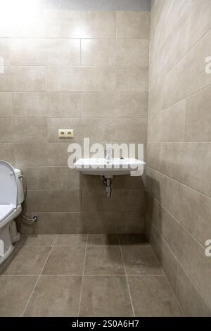 Une salle de bains minimaliste présente des murs et un sol carrelés beiges, un lavabo mural blanc avec un robinet simple et une partie visible d'une toilette. Banque D'Images