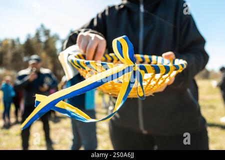 Une personne tient un panier tissé avec des rubans bleus et jaunes. En arrière-plan, les gens se rassemblent dans un espace naturel avec des arbres et un espace ouvert sous un clair Banque D'Images