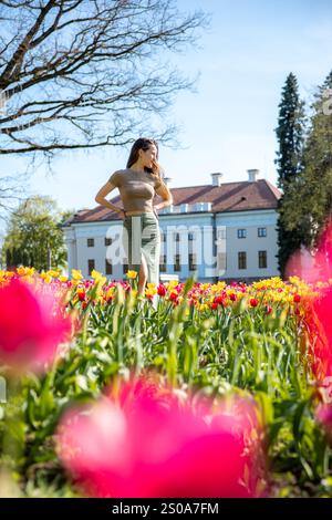 Une femme se tient dans un champ de tulipes animé, avec un bâtiment historique avec une façade bleu clair et un toit de tuiles rouges. De grands arbres encadrent la scène. Banque D'Images