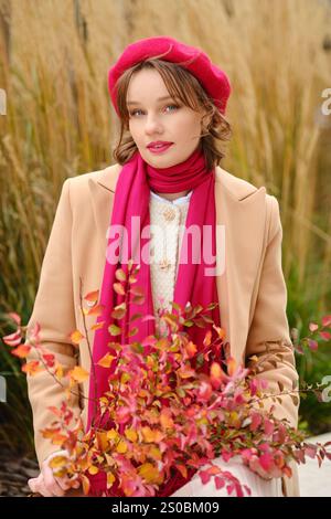 Une jeune femme se tient debout dans un parc orné d'un manteau chaud, d'un béret rouge et d'une écharpe, tenant des feuilles d'automne vibrantes. La toile de fond dorée de l'herbe ajoute au c Banque D'Images