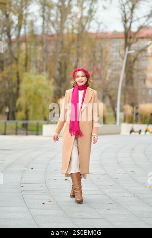 Une femme se promène dans un parc pittoresque en automne, vêtue d'un manteau beige, d'une écharpe et de bottes élégantes. Son expression joyeuse complète le vibra Banque D'Images