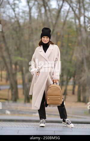 Une femme à la mode se tient en toute confiance dans un manteau léger et un bonnet noir, tenant un sac à dos élégant. La toile de fond présente une forêt sereine avec faible Banque D'Images