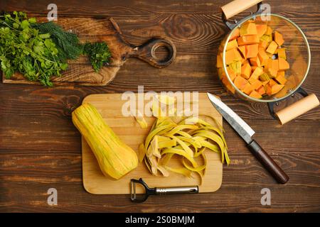 La courge butternut fraîche est pelée et hachée sur une planche à découper en bois, accompagnée d'herbes vibrantes et d'outils de cuisine bien rangés. L'ambiance est bri Banque D'Images