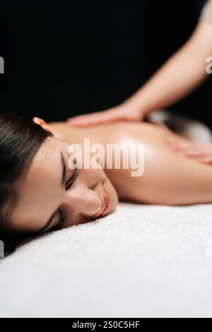 Gros plan sur le visage de la femme calme client se relaxant pendant le massage du dos dans le salon de spa avec intérieur sombre. Massothérapeute professionnel fournissant des soins thérapeutiques Banque D'Images