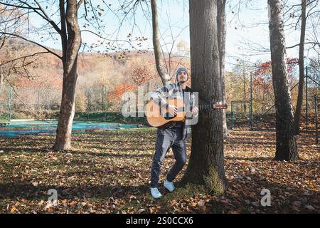 Homme portant une chemise à carreaux et un bonnet se tient dans un parc d'automne serein, caressant une guitare acoustique. Entouré de feuilles colorées et de grands arbres, il Banque D'Images