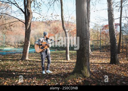 Homme portant une chemise à carreaux et un bonnet se tient dans un parc d'automne serein, caressant une guitare acoustique. Entouré de feuilles colorées et de grands arbres, il Banque D'Images