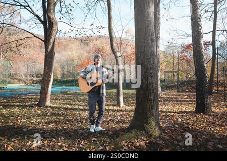 Homme portant une chemise à carreaux et un bonnet se tient dans un parc d'automne serein, caressant une guitare acoustique. Entouré de feuilles colorées et de grands arbres, il Banque D'Images