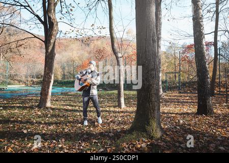 Homme portant une chemise à carreaux et un bonnet se tient dans un parc d'automne serein, caressant une guitare acoustique. Entouré de feuilles colorées et de grands arbres, il Banque D'Images