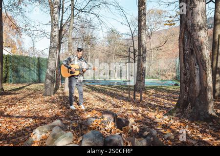 Guitariste dans une chemise à carreaux se tient confiant sur un tas de pierres dans un parc d'automne, caressant sa guitare acoustique. Entouré de feuilles dorées et ta Banque D'Images