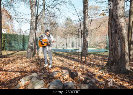 Guitariste dans une chemise à carreaux se tient confiant sur un tas de pierres dans un parc d'automne, caressant sa guitare acoustique. Entouré de feuilles dorées et ta Banque D'Images