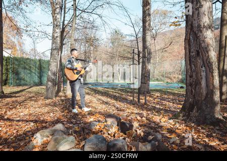 Guitariste dans une chemise à carreaux se tient confiant sur un tas de pierres dans un parc d'automne, caressant sa guitare acoustique. Entouré de feuilles dorées et ta Banque D'Images