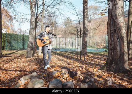 Guitariste dans une chemise à carreaux se tient confiant sur un tas de pierres dans un parc d'automne, caressant sa guitare acoustique. Entouré de feuilles dorées et ta Banque D'Images