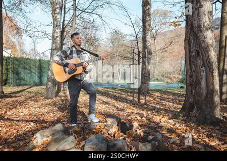 Guitariste dans une chemise à carreaux se tient confiant sur un tas de pierres dans un parc d'automne, caressant sa guitare acoustique. Entouré de feuilles dorées et ta Banque D'Images