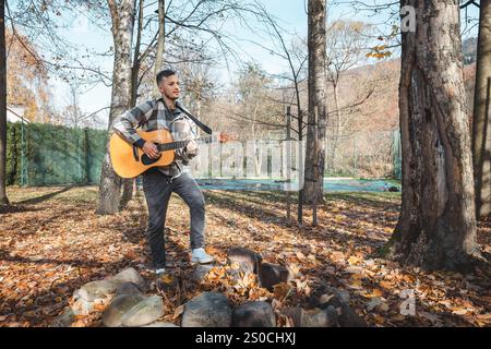 Guitariste dans une chemise à carreaux se tient confiant sur un tas de pierres dans un parc d'automne, caressant sa guitare acoustique. Entouré de feuilles dorées et ta Banque D'Images