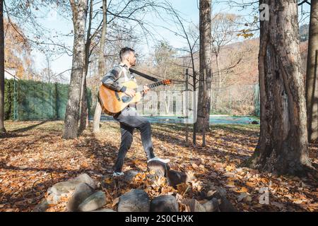 Guitariste dans une chemise à carreaux se tient confiant sur un tas de pierres dans un parc d'automne, caressant sa guitare acoustique. Entouré de feuilles dorées et ta Banque D'Images