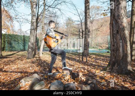 Guitariste dans une chemise à carreaux se tient confiant sur un tas de pierres dans un parc d'automne, caressant sa guitare acoustique. Entouré de feuilles dorées et ta Banque D'Images