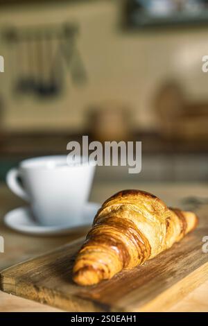 Tasse de café avec croissant sur une table de cuisine rustique. Croissants frais au premier plan sur planche de bois. Petit déjeuner à la maison, lumière chaude et atmosph confortable Banque D'Images