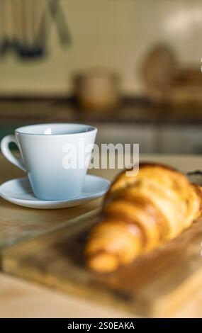 Tasse de café avec croissant sur une table de cuisine rustique. Croissants frais au premier plan hors de l'accent sur la planche de bois. Petit déjeuner à la maison, lumière chaude et Banque D'Images