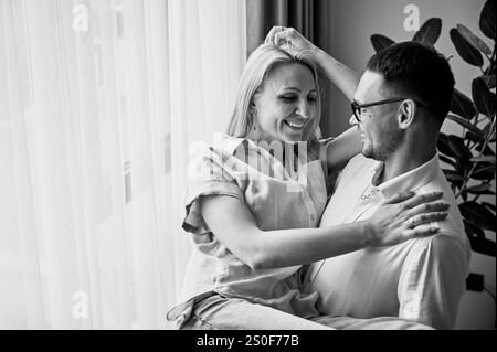 Homme heureux tenant sa femme dans ses bras à la maison. Charmant couple souriant l'un à l'autre. Doux couple se regardant dans des vêtements décontractés sur fond clair. Image en noir et blanc. Banque D'Images