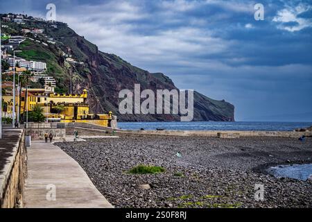 Une vue pittoresque sur un sentier côtier en bord de mer, avec des bâtiments en bord de mer, des falaises luxuriantes et une plage de galets sous un ciel nuageux serein. Le lo Banque D'Images