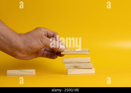 Une image montrant un homme plaçant des blocs de bois Jenga sur trois blocs de bois disposés soigneusement sur un fond jaune Uni. Banque D'Images