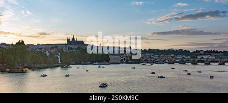 Une vue panoramique de Prague au coucher du soleil, avec l'emblématique château de Prague et le pont Charles sur la rivière Vltava. On voit des bateaux sur l'eau, avec un BE Banque D'Images