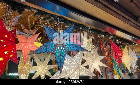 Assortiment de lanternes étoiles en papier illuminées de différentes couleurs et motifs suspendus à une structure en bois, décorés de guirlande à feuilles persistantes. Banque D'Images