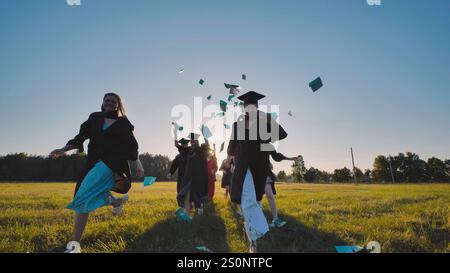 Des diplômés joyeux célébrant leur graduation en jetant des diplômes et des mortarboards dans l'air dans un champ de coucher du soleil Banque D'Images