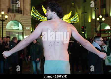 Londres, Royaume-Uni. 28 décembre 2024. Un homme vêtu uniquement de caleçons et portant « Doctors 4 Gaza » écrit sur son dos se tient debout dans la nuit froide à Piccadilly Circus alors que des partisans de la Palestine organisent une veillée d'urgence pour Gaza suite à un assaut israélien contre l'hôpital Kamal Adwan dans le nord de Gaza, l'un des derniers établissements de santé encore debout, et l'expulsion forcée de patients et du personnel médical. Les manifestants ont appelé à un cessez-le-feu, à une aide médicale urgente et à la responsabilité des violations du droit international. Crédit : Ron Fassbender/Alamy Live News Banque D'Images