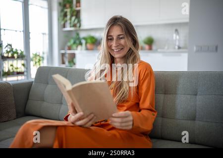 Femme heureuse se prélasse sur le canapé, absorbé dans le livre de lecture avec le sourire de contenu. Temps à la maison pour les amoureux des livres Banque D'Images
