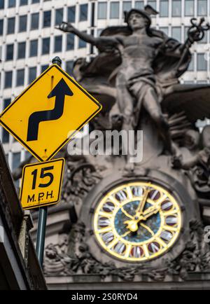 Gros plan d'un panneau routier jaune indiquant un virage serré devant vous, avec une limite de vitesse de 15 MPH. En arrière-plan, une statue détaillée et une grande horloge A. Banque D'Images