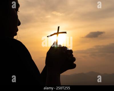 Silhouette d'une femme chrétienne tenant une croix priant au coucher du soleil. Femme réfléchie pliant les mains dans un geste de prière. Le concept de foi en Dieu Banque D'Images