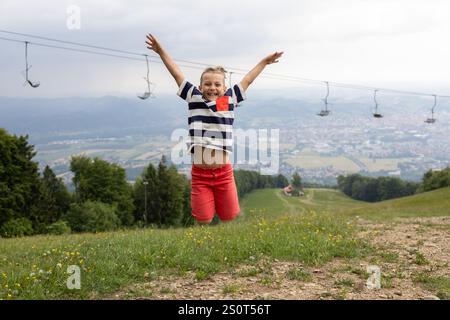 Un garçon de 8 ans saute joyeusement et crie de joie tout en marchant sur une montagne au-dessus de la ville. Concept d'enfance heureuse. Randonnée joyeuse et tra Banque D'Images