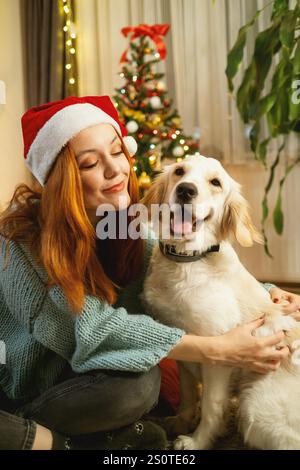Une jeune femme portant un chapeau de Père Noël embrasse amoureusement son Golden retriever tout en étant assise sur le sol, avec un sapin de Noël joliment décoré dans la BA Banque D'Images