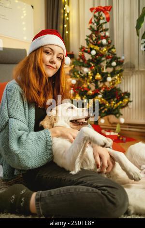 Une jeune femme portant un chapeau de Père Noël embrasse amoureusement son Golden retriever tout en étant assise sur le sol, avec un sapin de Noël joliment décoré dans la BA Banque D'Images