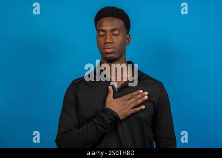 Portrait studio d'un jeune homme noir avec un pendentif africa, main sur la poitrine, les yeux fermés, sur un fond bleu vif Banque D'Images