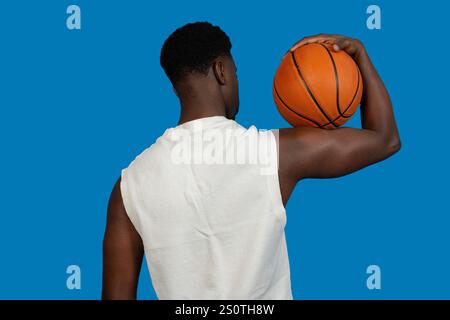 Joueur de basket-ball tient un ballon de basket-ball sur son épaule, vu de dos, sur un fond bleu vif Banque D'Images