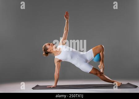 Jeune femme en vêtements de sport blancs pratiquant le yoga et s'étirant avec une petite balle en studio. Femme athlétique assise sur le tapis faisant la pose de yoga. Banque D'Images
