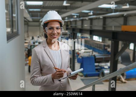 Jeune femme indienne, directrice d'usine métallurgique réussie écrivant des notes après inspection de production, portrait souriant. Banque D'Images