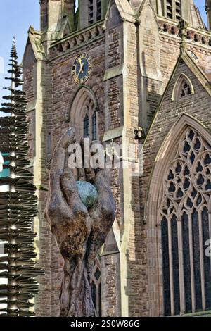 Une sculpture en bronze de mains berçant une sphère se tient bien en évidence au premier plan, avec une grande église en pierre formant le fond flou. La scul Banque D'Images