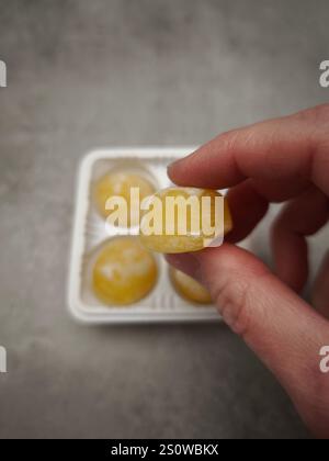Ramasser à la main un seul mochi à la mangue jaune dans un plateau en plastique, prêt à profiter d'une friandise sucrée Banque D'Images